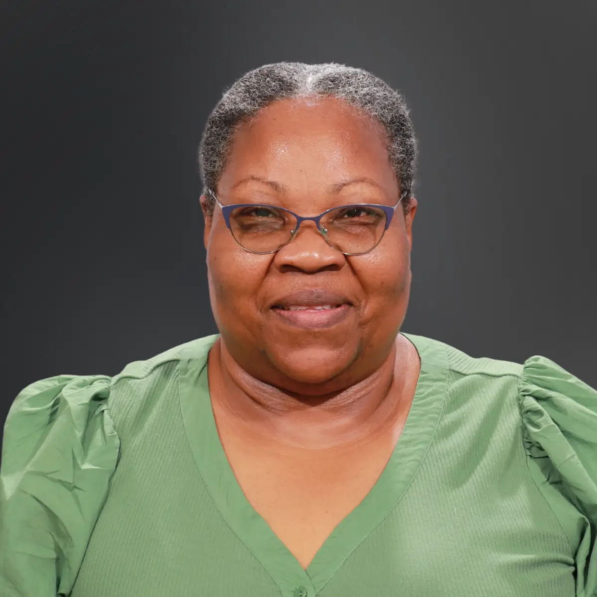 Portrait of a smiling Black woman with short gray hair, blue glasses, and a green blouse against a dark gray background.